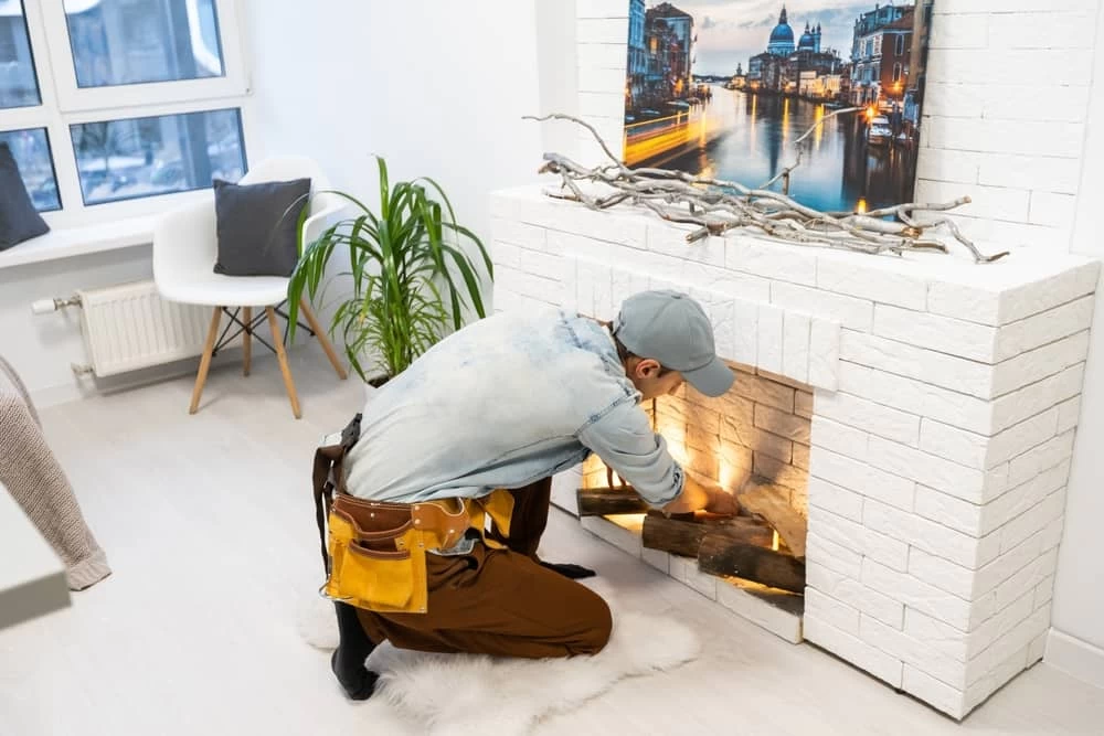 A person wearing a cap and tool belt is kneeling in front of a white brick fireplace, carefully placing logs inside after a thorough chimney sweep. There is a framed picture of a cityscape above the mantel, a potted plant, and a window in the background.