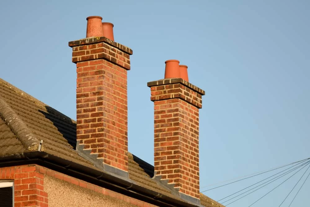 Two red brick chimneys with terracotta pots atop a roof, set against a clear blue sky, stand as part of a traditional house. One side of the roof is visible, highlighting the need for chimney repair.