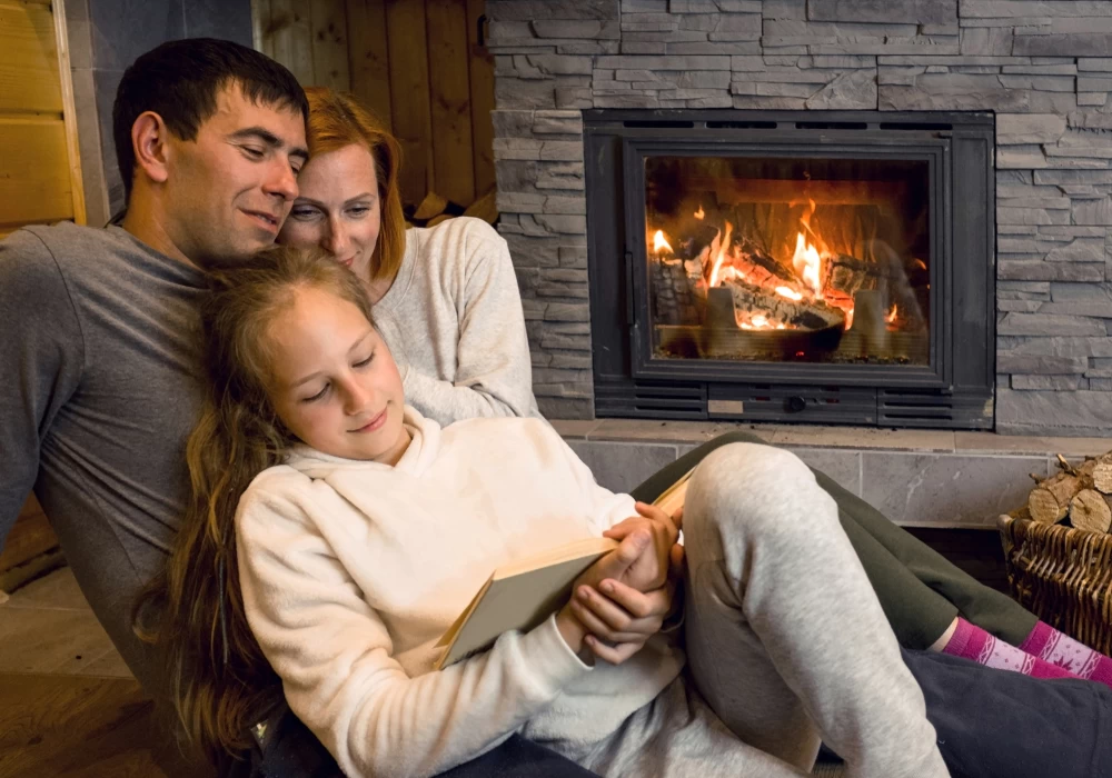 A family of three sits cozily by a lit fireplace. A woman and a man cuddle behind a girl who is seated in front, reading a book.
