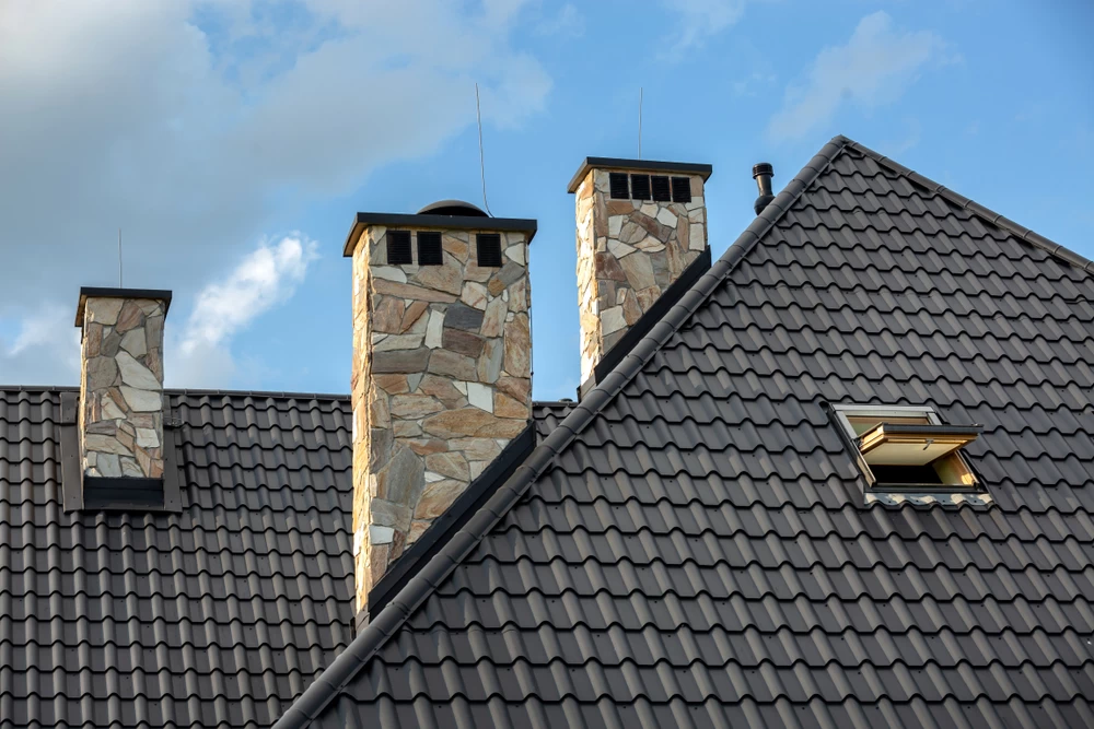 A rooftop with three stone chimneys and a tiled surface under a blue sky. One chimney has a metal cap, and there is a skylight window on the sloped roof.
