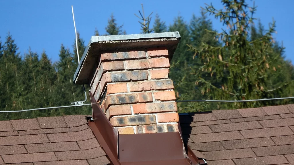 A brick chimney on a sloped roof appears bent, with a metal cap on top. The roof is covered in brown shingles, and trees can be seen in the background against a clear blue sky.