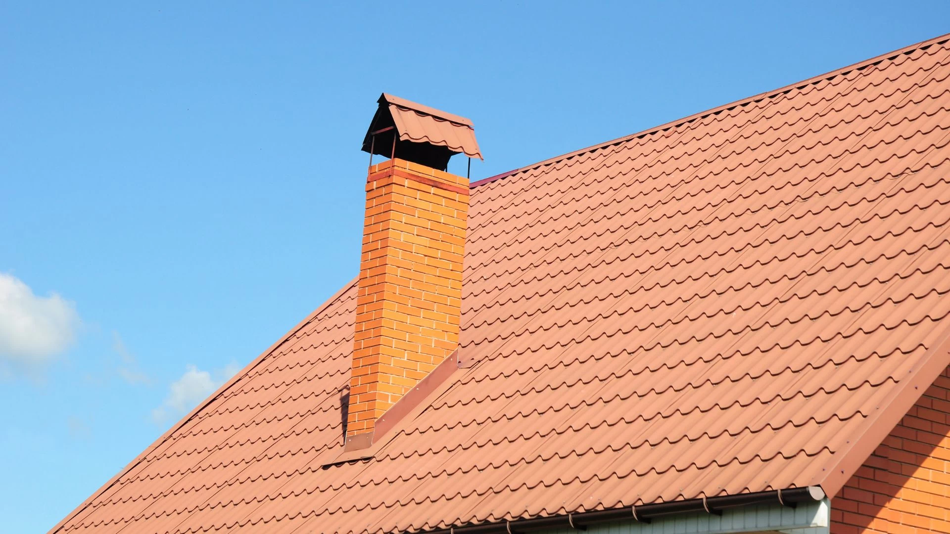 Red brick chimney on a house with a red metal roof against a clear blue sky.