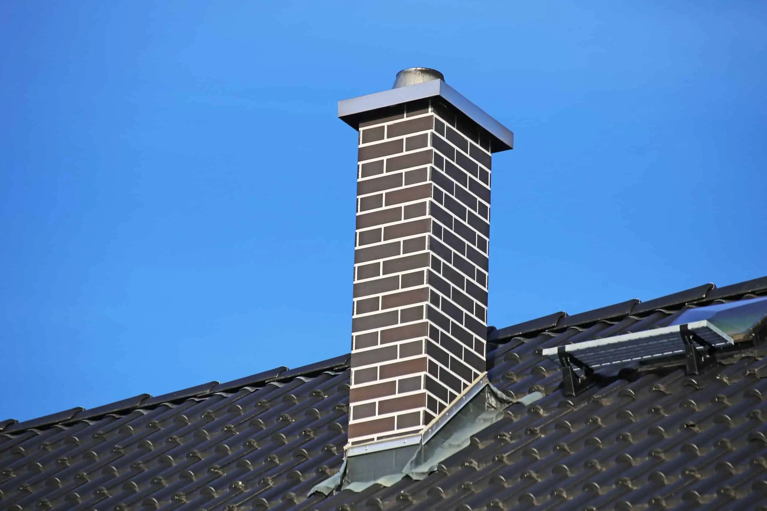A brick chimney with a metal cap on a tiled roof under a clear blue sky.