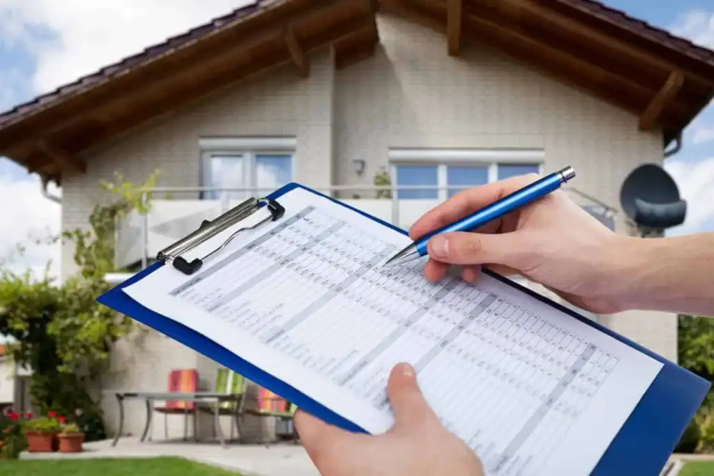 A person holding a blue clipboard with a checklist or inspection form in front of a modern house with a sloped roof and outdoor furniture.